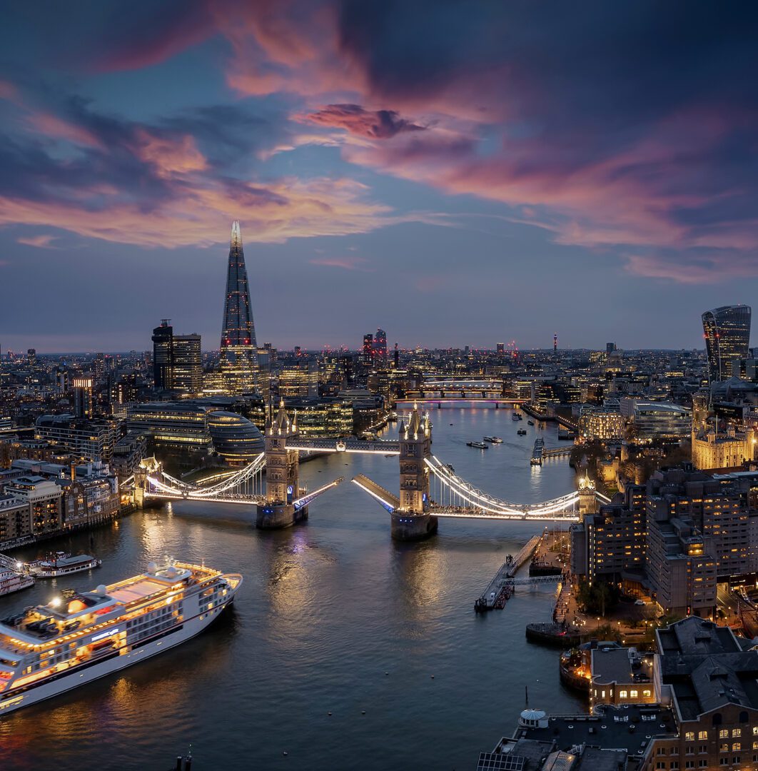 Panoramic, aerial view of the skyline of London with a motion blurred cruise ship passing under the lifted Tower Bridge during dusk, England